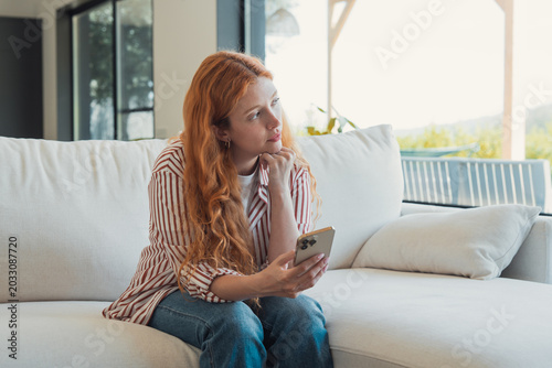 Fotografie Thoughtful young adult woman with red hair sitting on a white sofa, holding a mo