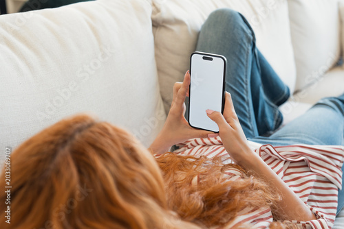 Redhead woman lounging on a white sofa at home, holding a modern smartphone with a blank white screen, scrolling and tapping while relaxing in daylight, overhead crop