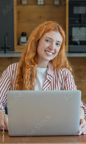 Smiling woman with red hair looking at the camera while working remotely on her laptop, depicting modern work flexibility, career success, and a connected lifestyle