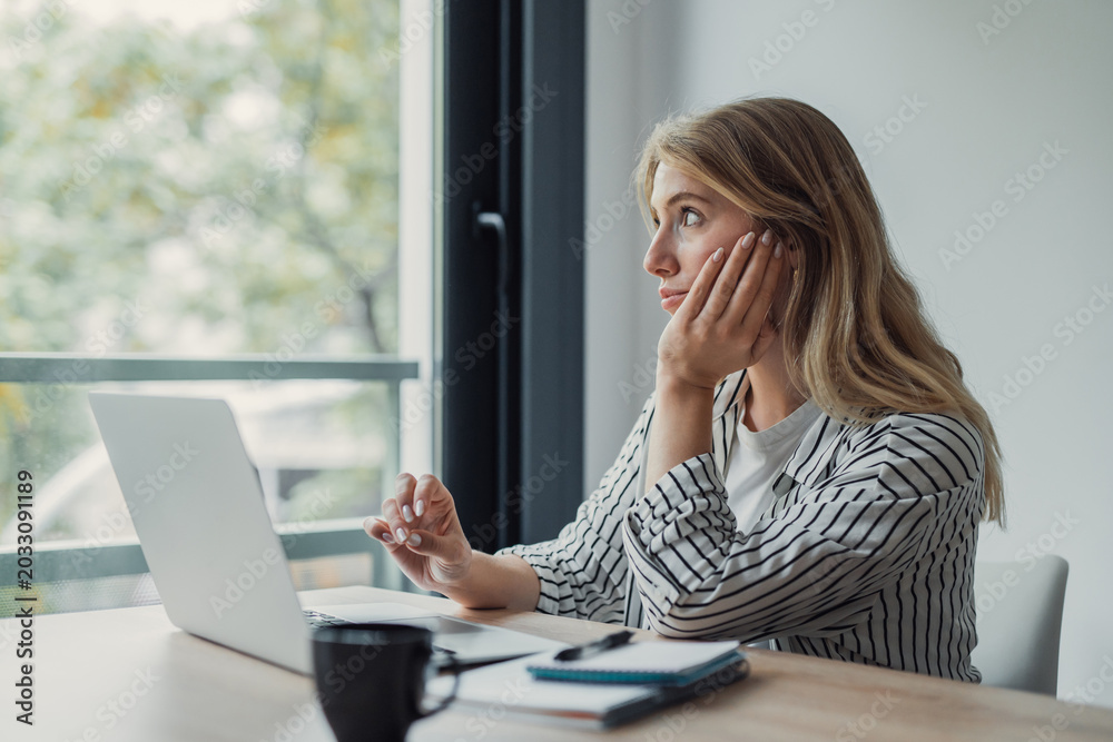 Fototapeta premium Young woman looking away from her laptop, sitting at a desk and contemplating with her hand resting on her face, possibly taking a break from remote work
