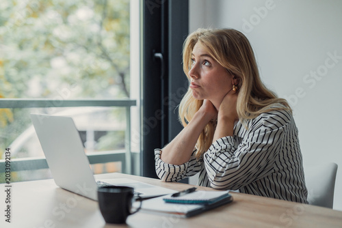 Quadro em tela Young adult woman appearing tired and unmotivated, sitting at a desk with a lapt