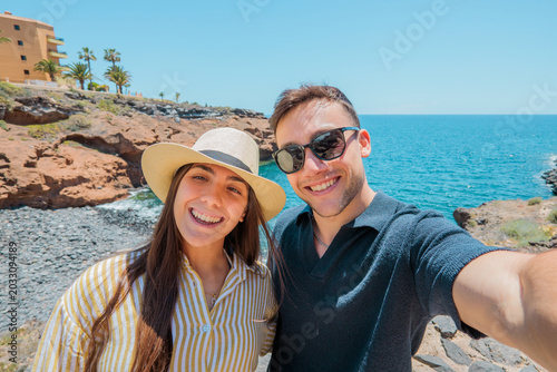 Young couple capturing a joyful selfie moment during their summer vacation, standing near the rocky coast with the blue ocean and clear sky in the background, embracing travel and adventure
