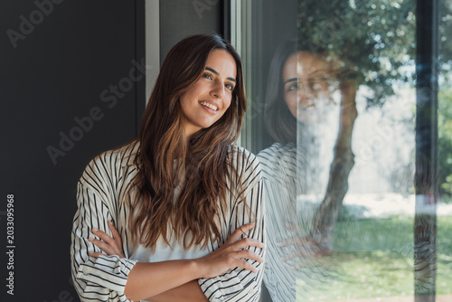 Young contented woman smiling with arms crossed leaning against a window, reflecting lush green garden, representing concepts of contemplation, future, home, well-being, and nature connection