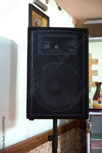 stone wall background, perfect for events, concerts, parties and public speaking setups. Professional large black audio speaker mounted on a sturdy metal tripod stand indoors with a wooden floor .