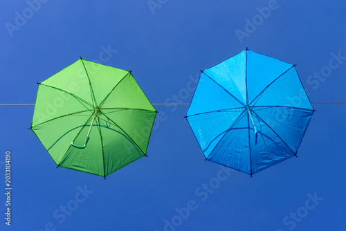 two colourful umbrellas, one green and one blue, hanging from a rope against a backdrop of blue sky on a sunny day
