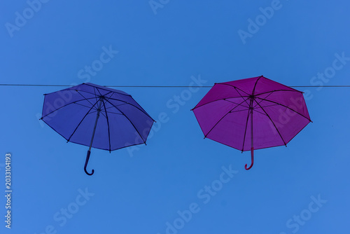 two colourful umbrellas, one purple and one navy blue, hanging from a rope against a backdrop of blue sky on a sunny day