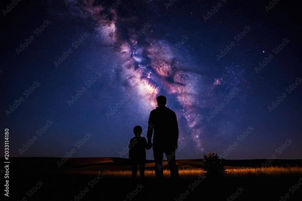 Fototapeta premium Father and child hold hands watching starry Milky Way galaxy above dark landscape. People gaze at night sky, learn astronomy, share family bonding time, dream together.