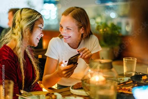 Two smiling young women sharing a phone at a cozy restaurant