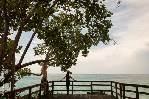 Canvas Print Serene tropical scene of a woman standing on a wooden oceanfront deck surrounded by lush coastal trees and overlooking the calm sea