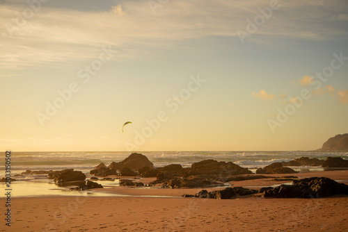 Golden sunset over the Atlantic Ocean with black volcanic stones scattered along the beach, while a kiteboarder rides in the distant horizon under a glowing sky, capturing the energy of wind and sea.