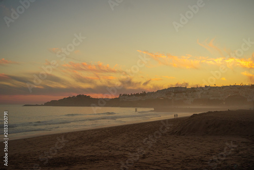 Golden sunset over a misty sandy beach in Portugal’s Algarve region, with soft pink clouds in the sky, a hazy ocean horizon, and distant coastal hills dotted with houses glowing in warm evening light.