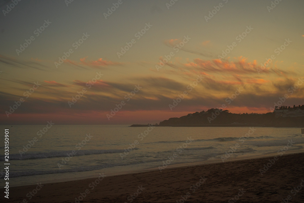 Obraz premium Golden sunset over a misty sandy beach in Portugal’s Algarve region, with soft pink clouds in the sky, a hazy ocean horizon, and distant coastal hills dotted with houses glowing in warm evening light.