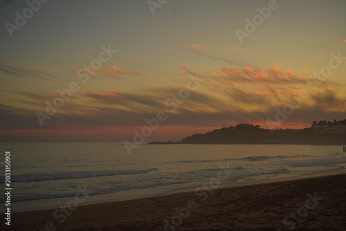 Golden sunset over a misty sandy beach in Portugal’s Algarve region, with soft pink clouds in the sky, a hazy ocean horizon, and distant coastal hills dotted with houses glowing in warm evening light.