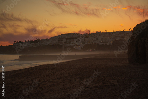 Golden sunset over a misty sandy beach in Portugal’s Algarve region, with soft pink clouds in the sky, a hazy ocean horizon, and distant coastal hills dotted with houses glowing in warm evening light.