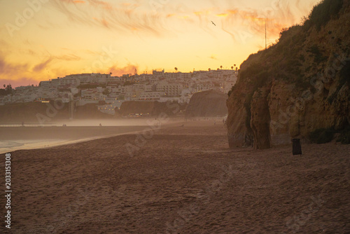 Golden sunset over a misty sandy beach in Portugal’s Algarve region, with soft pink clouds in the sky, a hazy ocean horizon, and distant coastal hills dotted with houses glowing in warm evening light.