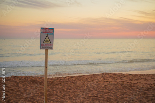 Warning sign on Algarve beach with Atlantic waves and glowing horizon of pink clouds, capturing contrast between human caution and wild coastal beauty.