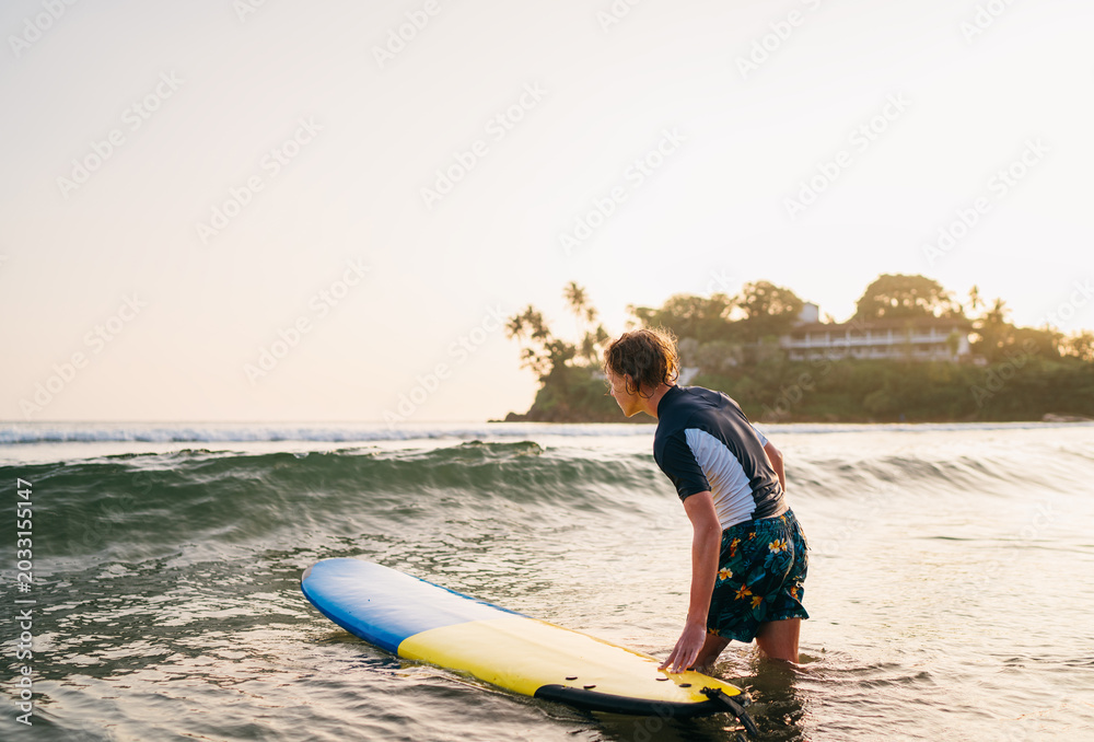 Fototapeta premium focused blonde teenage boy in rash guard holding surfboard while standing in ocean waves on Dewala Beach in Sri Lanka during sunset representing youth active lifestyle tropical travel adventure and wa