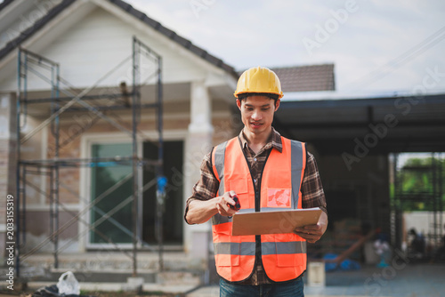 Young Asian construction worker or engineer checking building progress at a residential construction site, wearing hard hat and safety vest