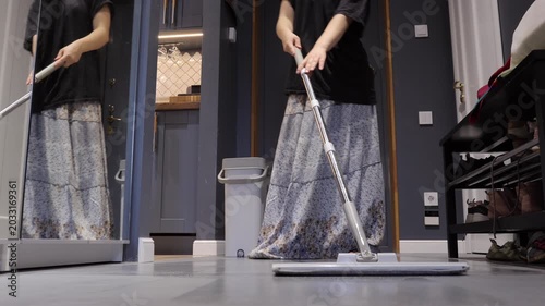 A teenager uses a flat mop near a mirrored closet and gray bucket while doing an entryway household chore.