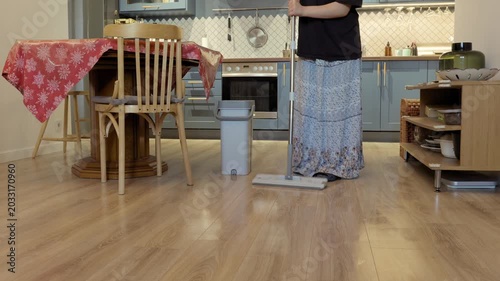 A teenager mops a light wooden floor near a dining table and gray bucket during indoor home cleaning.