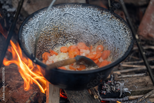 Vegetables simmering in an enamel pot over a campfire in the forest. Cozy outdoor cooking atmosphere featuring fire flames, wooden spoon, rustic style, and traditional camping experience.