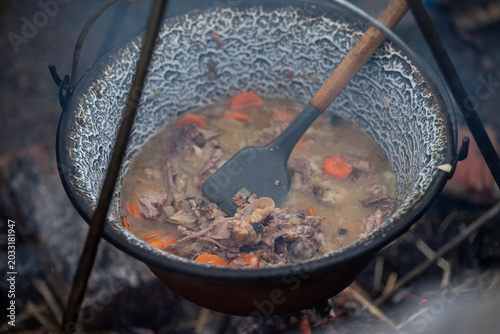 Close-up of carrots and onions being cooked in a hanging pot above an open fire in nature. Authentic camp cooking scene with rustic cookware, flames, smoke, and outdoor survival lifestyle concept.