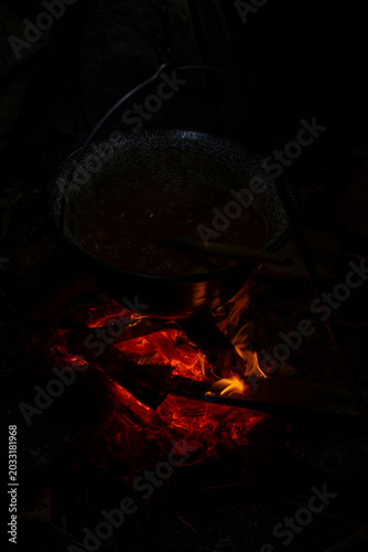 Cooking vegetables in a traditional enamel pot over an open campfire outdoors. Rustic outdoor cooking scene with carrots, onions, firewood, smoke, and camping atmosphere.