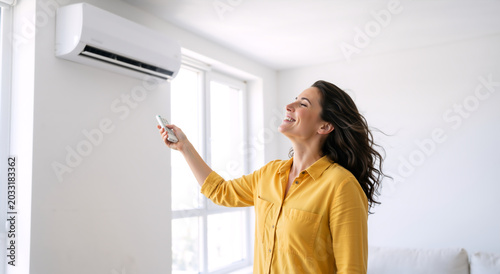 Smiling woman turning on air conditioner with remote control. Happy female enjoying cool breeze from ac unit at home in summer