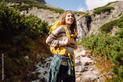 Female hiker with yellow backpack walks along mountain trail alongside stream. Woman enjoys  mountain scenery and feels a sense of freedom. Concepts of adventure, hiking, and sports.
