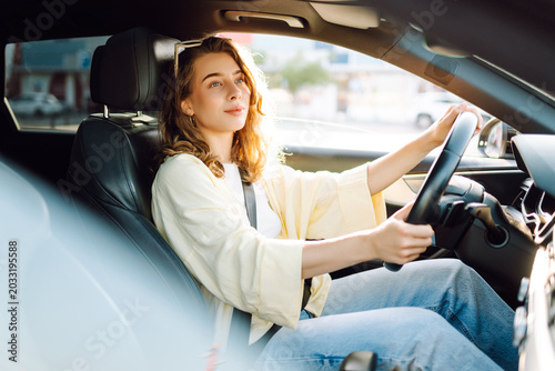 A young woman drives through a busy area during the day. She smiles, looking out the window. Sunlight floods the car's interior, creating a warm atmosphere. Urban and transportation concept.