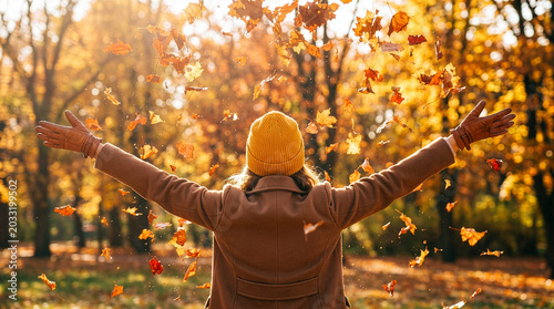 Fotografie Joyful woman in brown coat and yellow hat throwing autumn leaves in the air with
