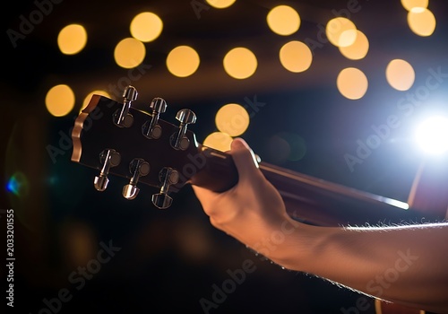 A musician's hand holding an acoustic guitar on an evening stage with warm bokeh light atmosphere