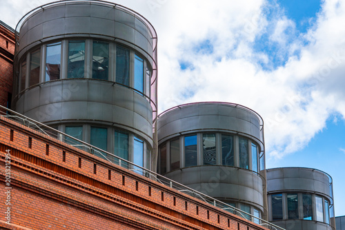 Tableau sur toile Modern cylindrical steel and glass structures rising above a historic red brick building facade against a blue sky with white clouds in bright daylight