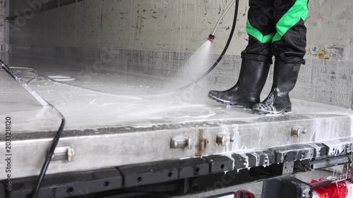 A uniformed cleaner washes the body of a large truck with a powerful jet of water with foam in rubber boots