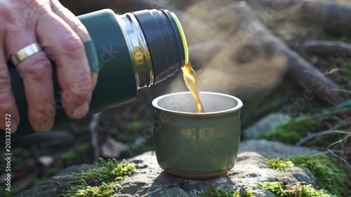 Stable ecu macro video of a human hand pouring rich, dark, steaming liquid from a forest-green thermos into an earthy green cup on a mossy rock. Bokeh background and rising steam. Serene warming