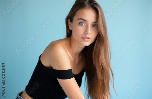 Young woman with long light brown hair looks directly ahead. She wears a black off shoulder top against soft blue background. Calm expression, composed posture, studio shot.