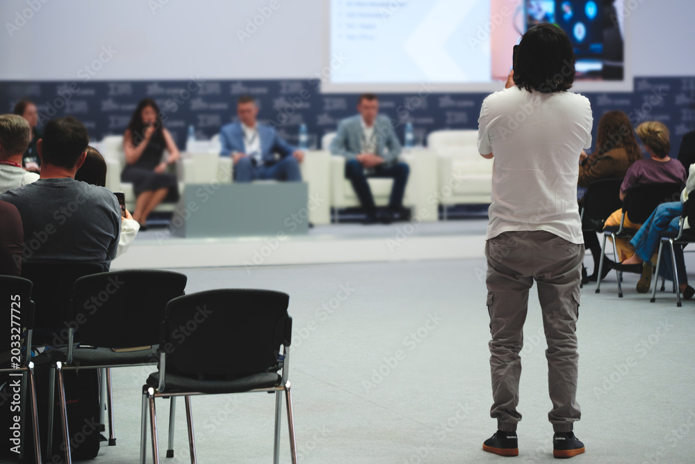 Fototapeta premium Person stands facing the conference panelists and records video on a smartphone. Moderator faces group of seated speakers. Audience watches attentively.