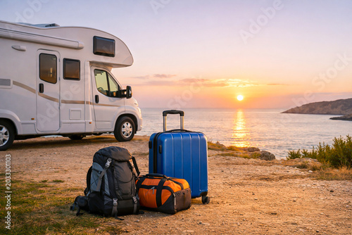 Luggage set with blue suitcase and backpacks near a motorhome parked on a coastal cliff at sunset