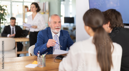 Elderly male bank employee at desk with laptop and discussing with young couple customers