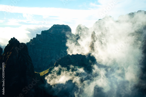 Fotografi Dramatic mountain crags and steep volcanic cliffs pierced by bright morning sunlight with rolling white clouds in the valley