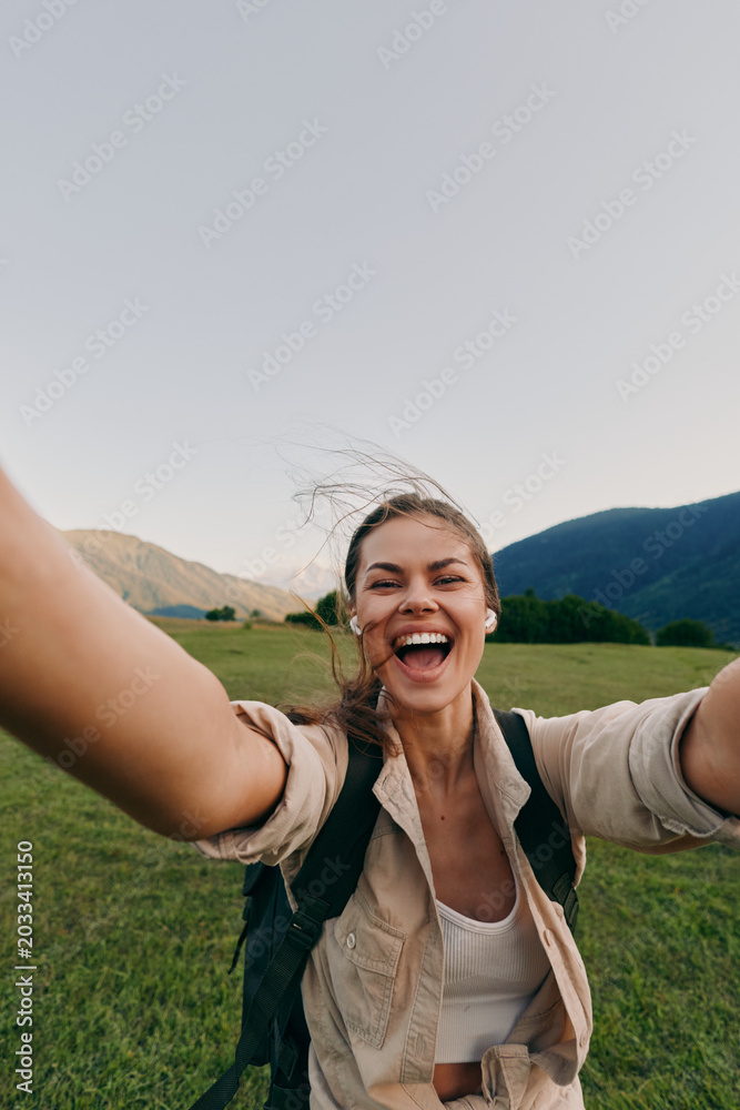 Fototapeta premium Vibrant woman outdoors, smiling with arms open, backpack ready, sunlit field and mountains behind, carefree lifestyle, candid moment, natural colors