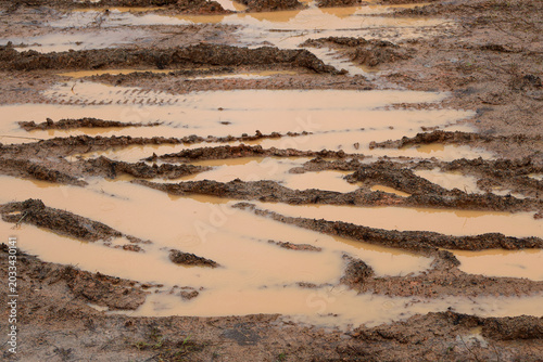 A high-angle shot of a dirty, wet dirt road filled with brown muddy water puddles and deep vehicle tire track marks after heavy rainfall.