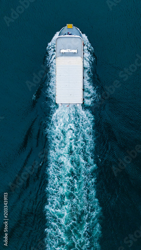 Aerial top down view of a small white motorboat cruising rapidly across deep dark blue ocean water leaving a prominent bright white foaming wake trail behind on the calm sea surface