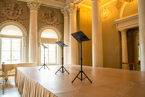 Opulent baroque concert hall interior with gilded column, arched window and black music stand on polished floor, luxury heritage venue concept.