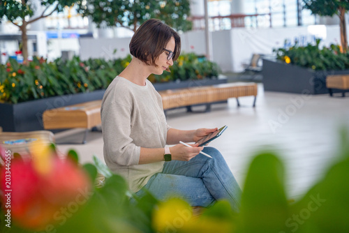 Cuadro en lienzo Young woman with glasses in beige sweater sitting on bench using tablet in bright modern indoor garden atrium with colorful flower in foreground, lifestyle remote work concept