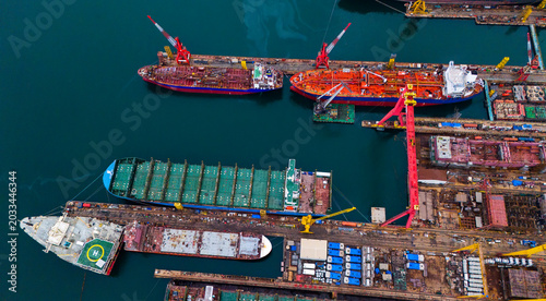 Aerial perspective of large industrial dockyard featuring multiple docked cargo vessels a green container ship massive red gantry cranes and deep blue ocean water