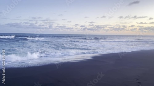 A serene view of ocean waves breaking on a dark sandy beach under a cloudy sky at dusk.
