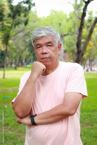 Portrait of a healthy Asian senior man smiling thoughtfully in a public park. Active retirement and elderly well-being concept.