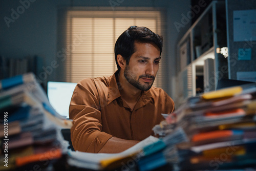 Overworked young Latin businessman working late on document in large stack of paperwork files at office night. Tax audit deadline, accounting stress, corporate burnout, urgent business concept.