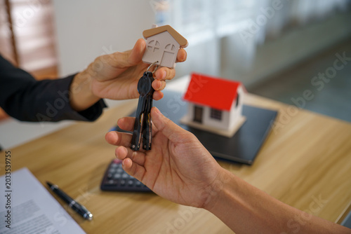 Real estate house key home buying key handover property sale mortgage closing as agent gives keys to buyer over contract on desk with house model calculator and pen joyful homeowner moment